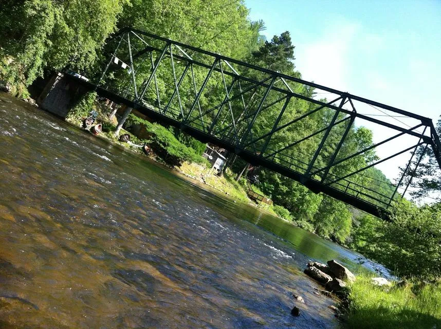 A metal truss bridge spans the flowing Toccoa river surrounded by lush green trees, with a house and grassy area visible in the background—an inviting scene from the best hikes in Blue Ridge, GA